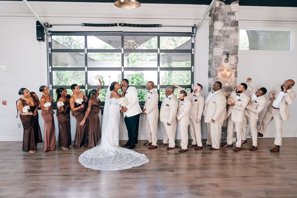 Bride and groom kiss at the center of a wedding party as bridesmaids in brown dresses and groomsmen in tan suits cheer in an indoor venue.