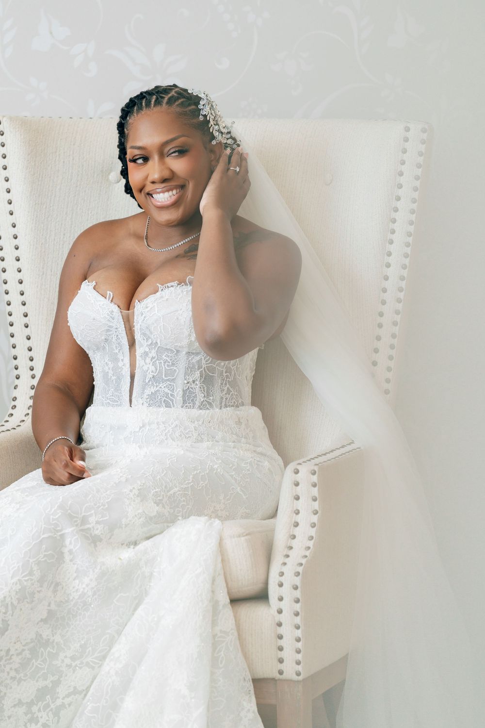 Radiant bride in a strapless lace wedding gown and long veil smiles while seated in a tufted white wing chair.