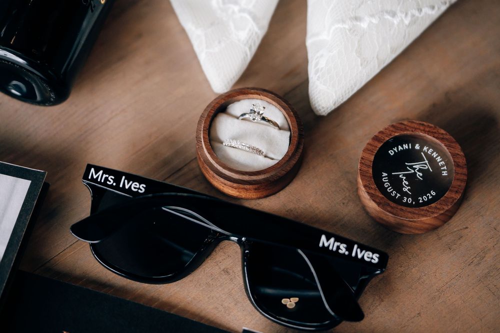 Wedding rings in a round wooden ring box beside black Mrs. Ives sunglasses and an engraved keepsake box on a rustic wooden table.