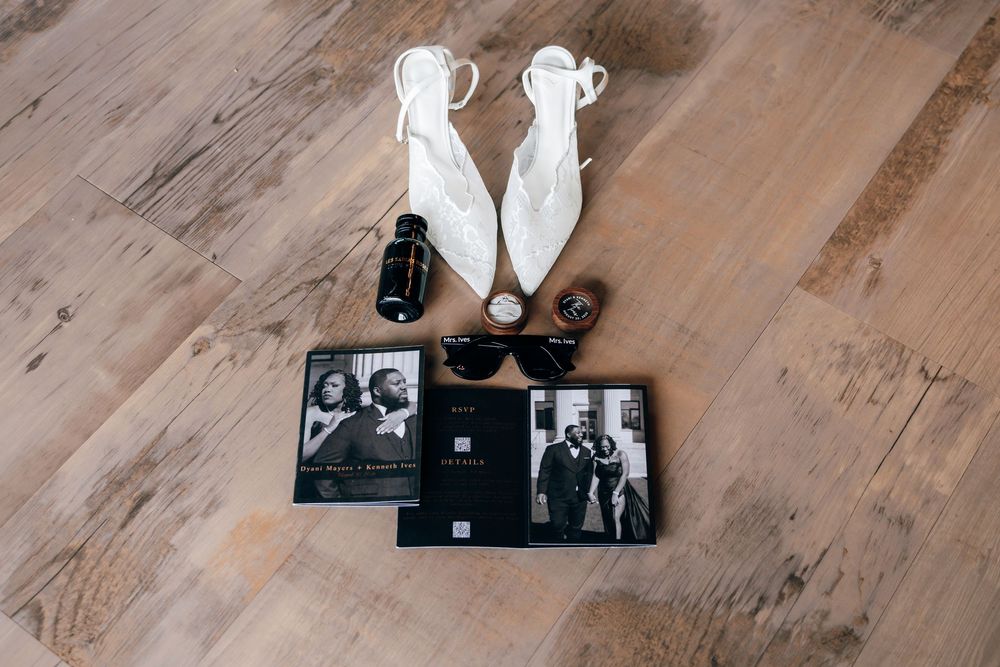 White pointed-toe wedding heels, black sunglasses, perfume bottle, rings and two black-and-white photo books on a rustic wood floor.