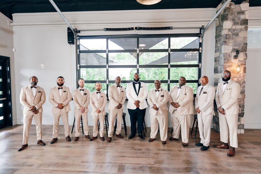Groom in a black tuxedo stands center among nine groomsmen in cream dinner jackets in a bright venue with large windows and a stone wall.