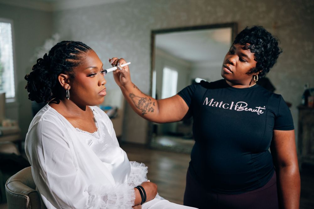 A makeup artist in a black t-shirt applies makeup to a woman in a white ruffled robe seated in a softly lit room with a mirror.
