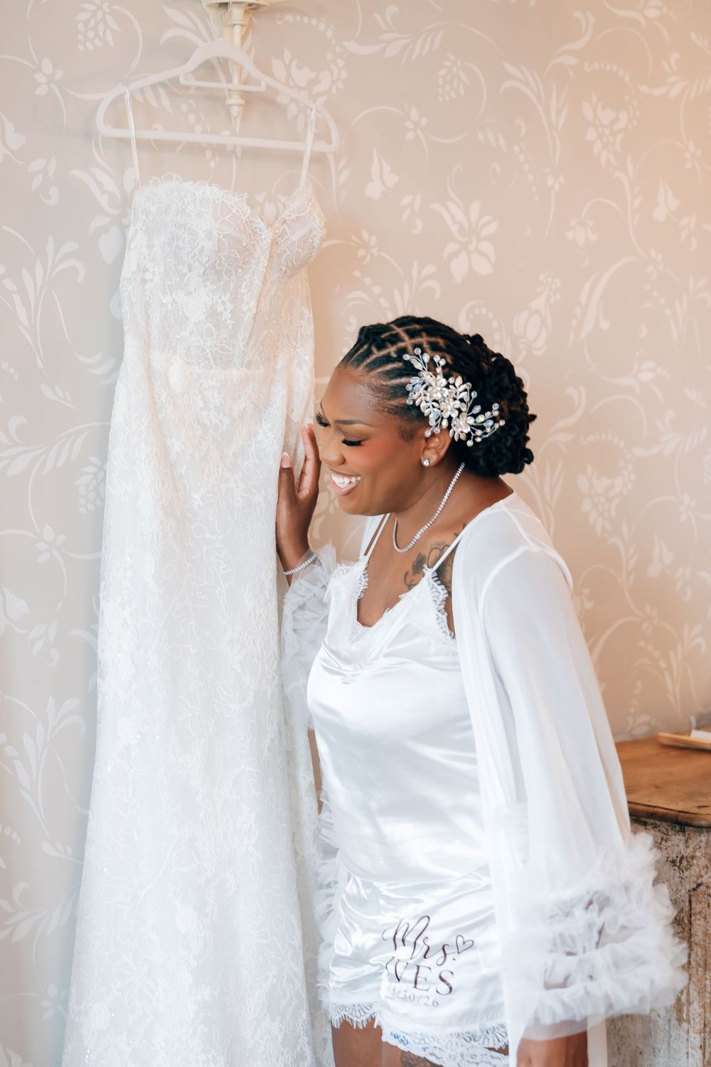 Smiling bride in a white satin robe and sparkling hair accessory leans against her lace wedding gown hanging on the wall.