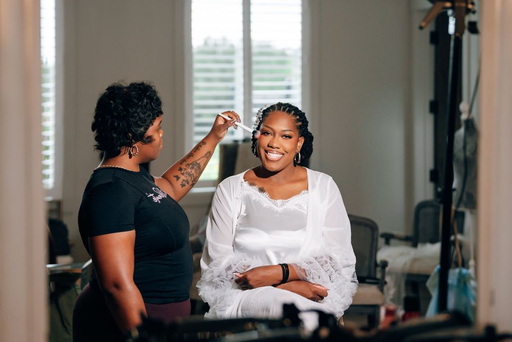 Smiling bride in a white feathered robe sits by a window while a makeup artist styles her hair during wedding day preparation.