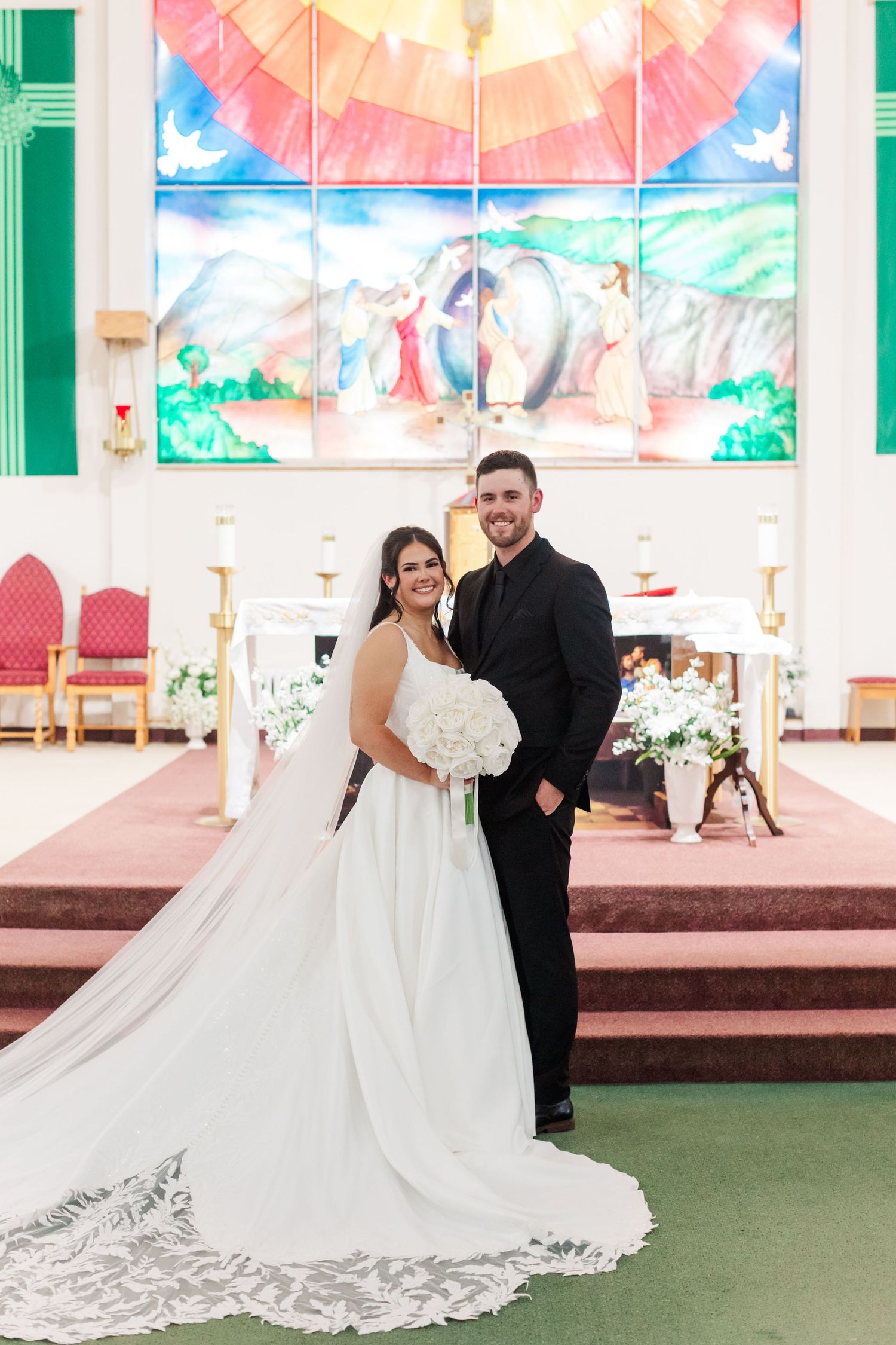 Bride in a white gown with a long lace veil holding a bouquet stands beside a groom in a black suit at a church altar.