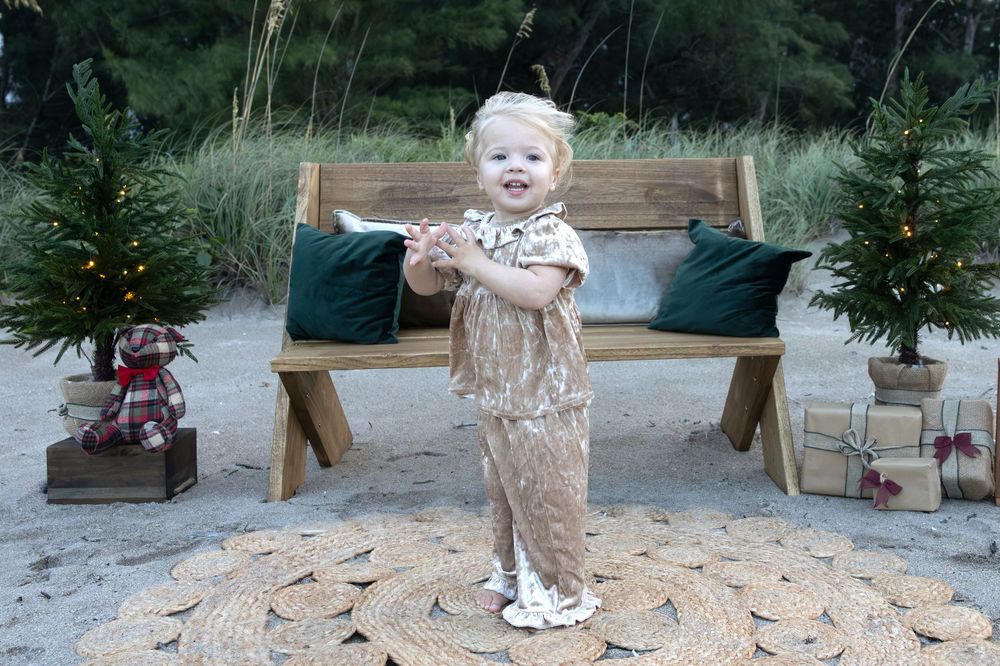 Toddler in a sparkly gold outfit stands on a woven mat at a beach holiday photo setup with a wooden bench, mini Christmas trees and gifts.