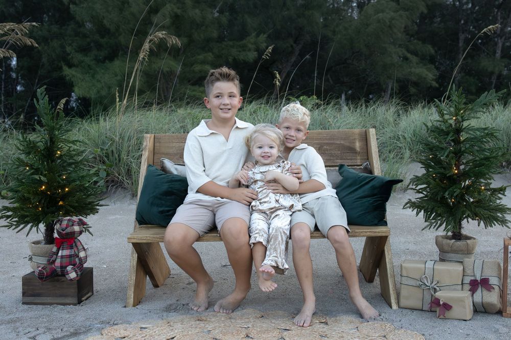 Two boys and a toddler sit together on a wooden bench on a beach, surrounded by small Christmas trees and wrapped holiday gifts.