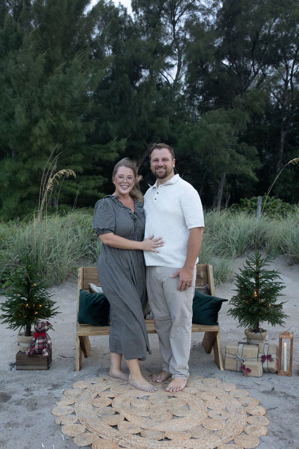 Smiling couple standing barefoot on a beach beside a wooden bench, mini Christmas trees, and wrapped gifts for a holiday portrait.