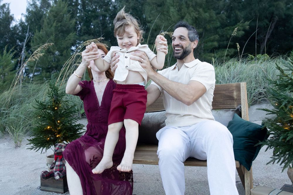 Smiling couple on a wooden beach bench lifting a laughing toddler in white and red outfit beside a small decorated Christmas tree.