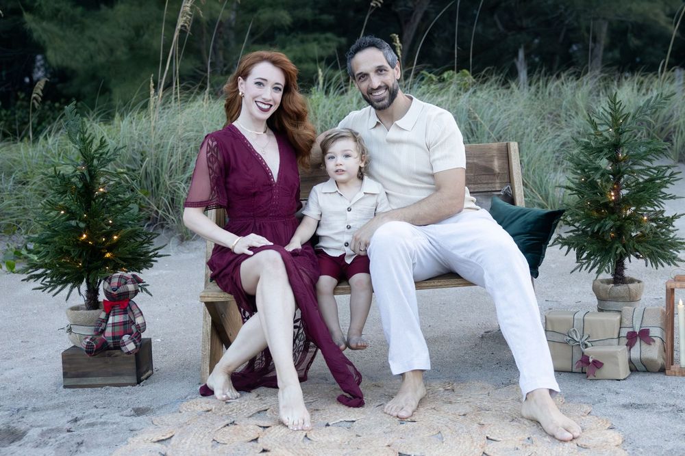 Family of three in a beach holiday portrait, a woman in a burgundy dress and a barefoot man in white sitting on a bench with a toddler.