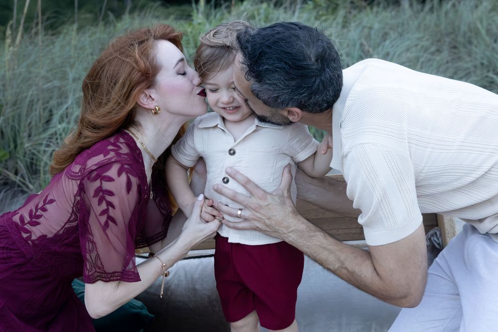 A red-haired woman in a burgundy lace dress and a man in a cream shirt kiss a smiling toddler in red shorts outdoors near tall grass.