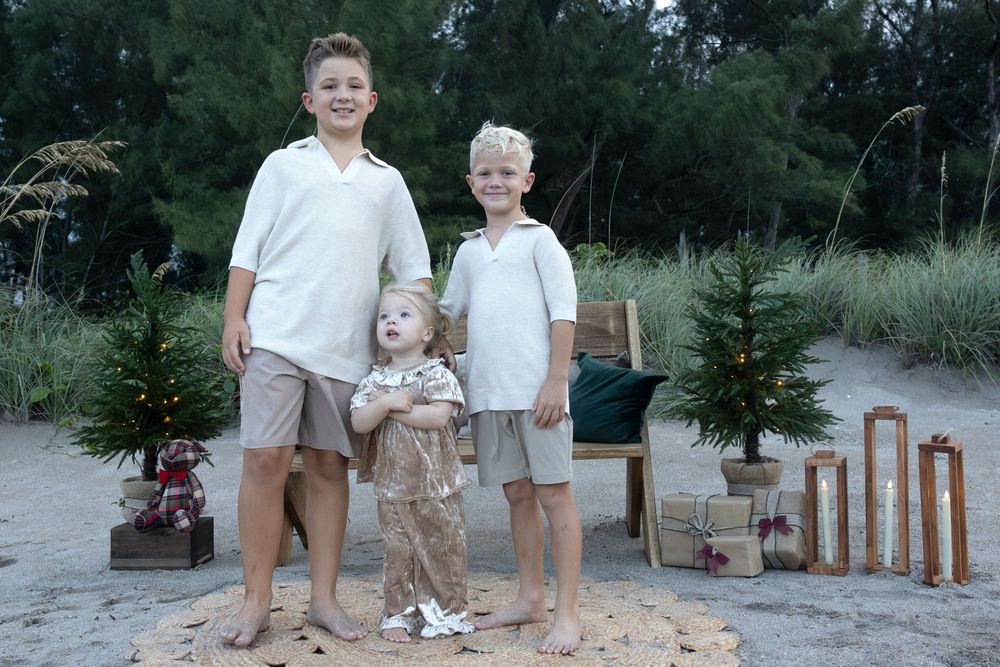 Two boys and a toddler pose barefoot on a beach beside small decorated Christmas trees, wrapped gifts, and a wooden chair at sunset.