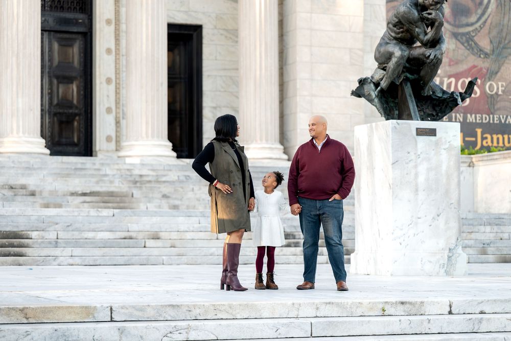 A family of three stands on marble steps outside a grand classical building with a bronze statue.