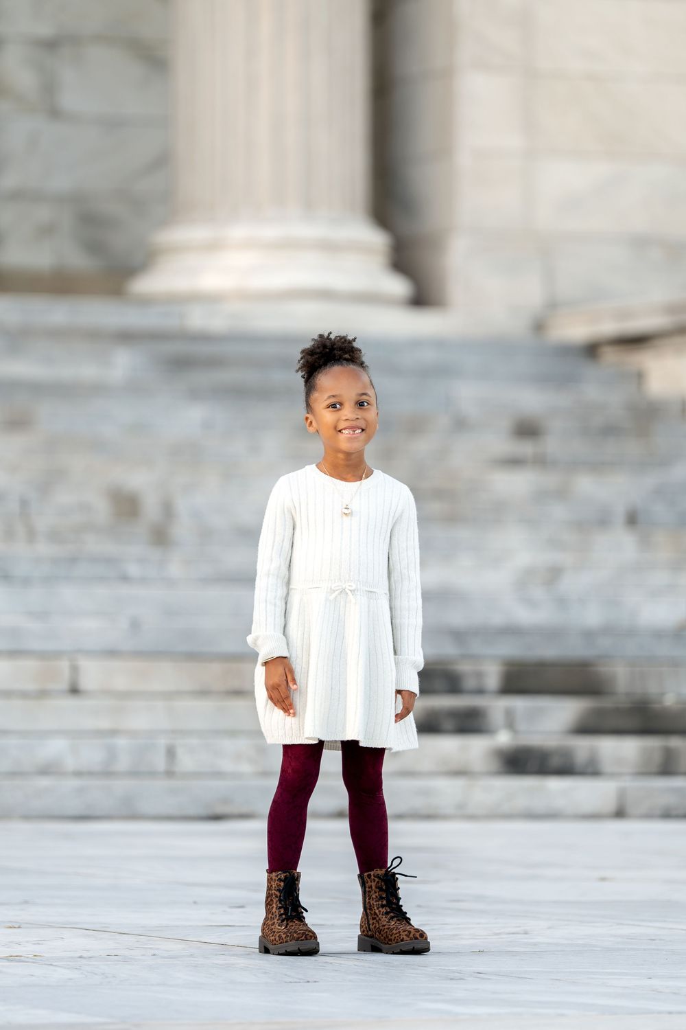 Young Black girl in white dress, burgundy tights, and boots smiling on marble steps.
