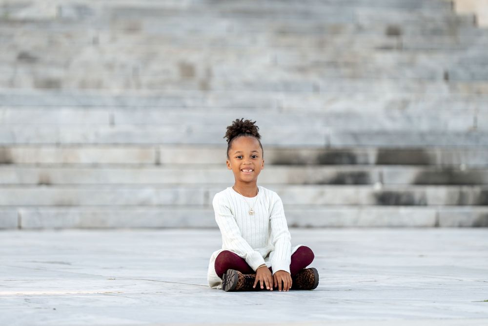 A smiling young Black girl sits cross-legged on marble steps wearing a white top and burgundy pants.