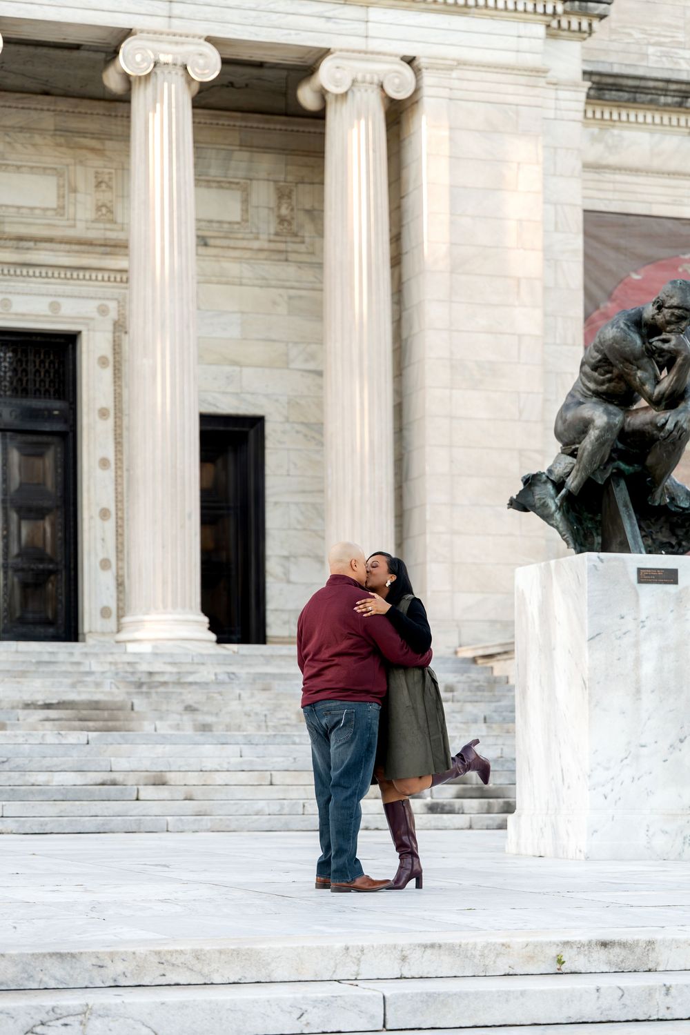 Couple sharing a romantic kiss on marble steps outside a neoclassical building with tall columns and a bronze statue.