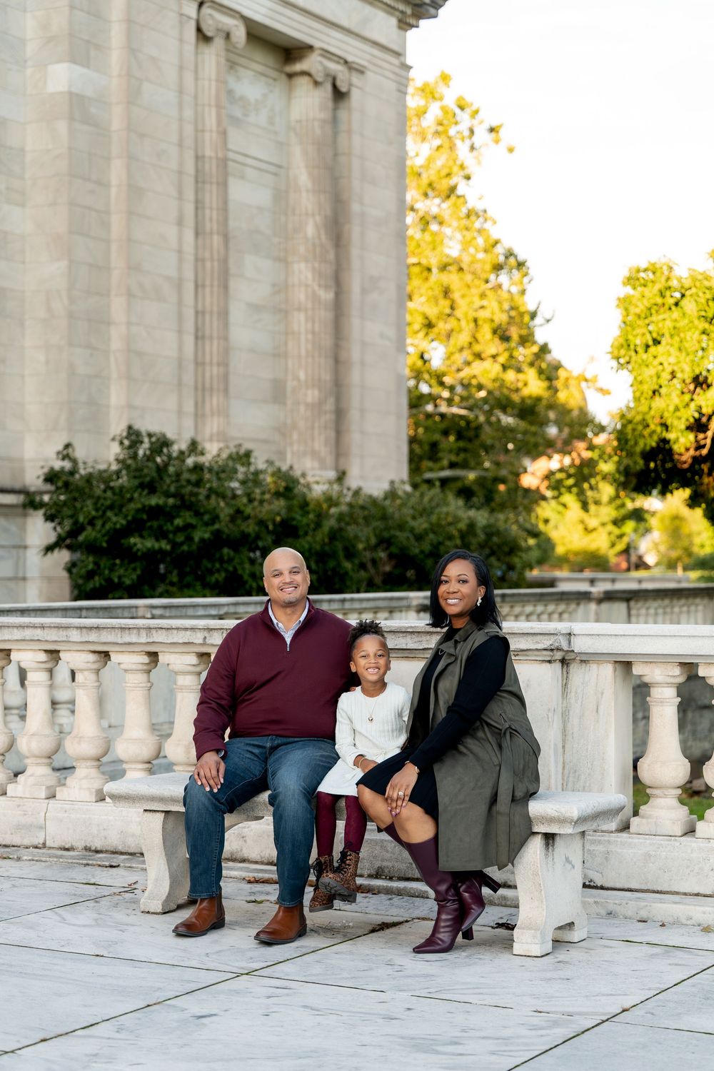 A family of three poses together on marble steps near a classical stone building outdoors.