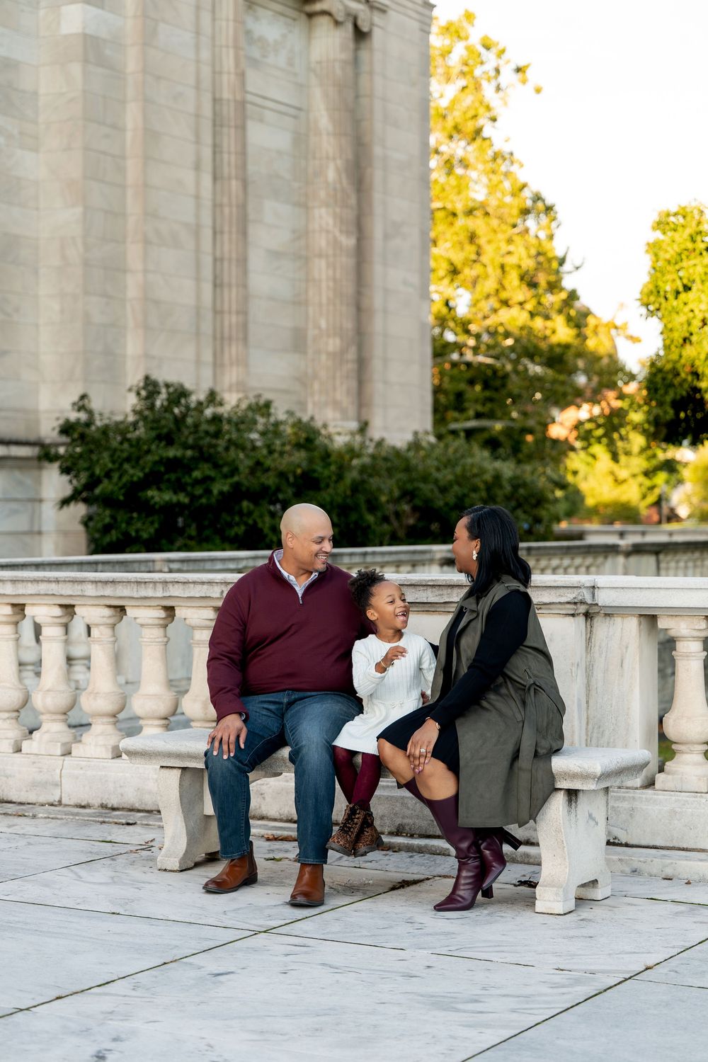 A family of three sits on a marble bench, smiling at each other near a classical building.