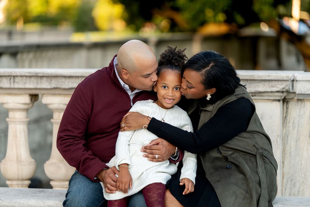 A loving Black family with parents kissing their young daughter on the cheeks outdoors near stone railings.