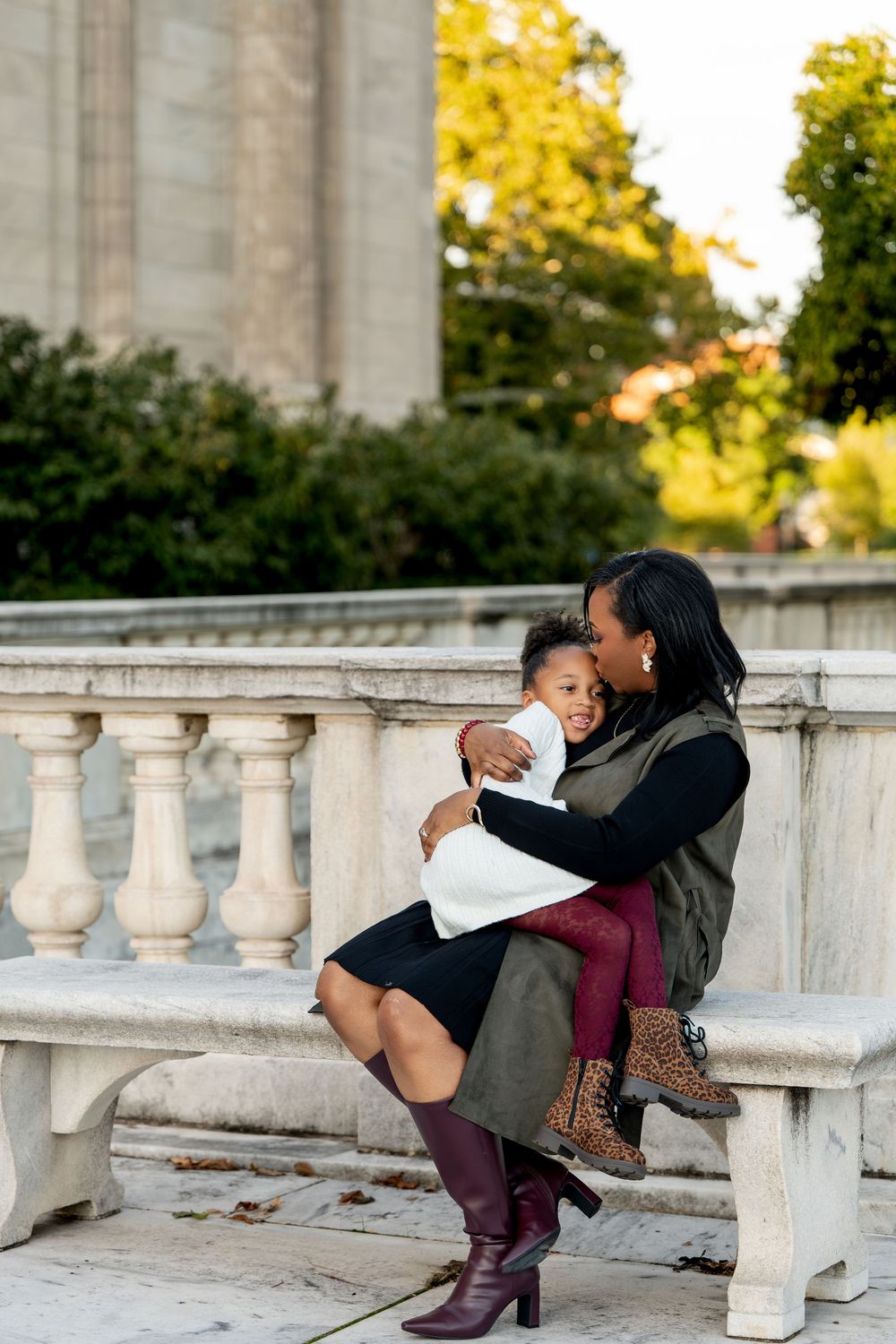 A mother sits on a stone bench outdoors, lovingly embracing her young daughter in a warm autumn setting.