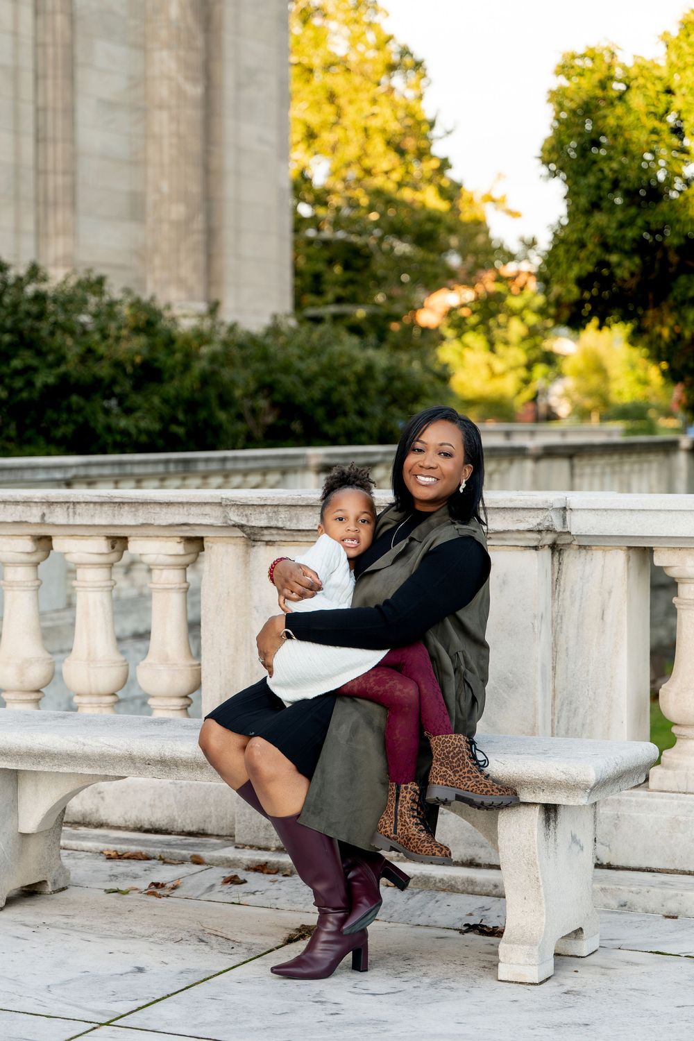 A smiling mother sits on a marble bench outdoors, lovingly holding her young daughter in her lap.