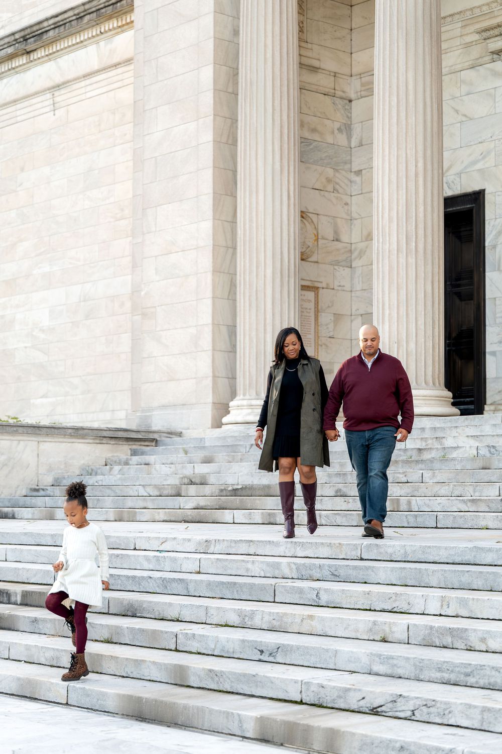 A family of three descends grand marble steps outside a neoclassical building with tall white columns.