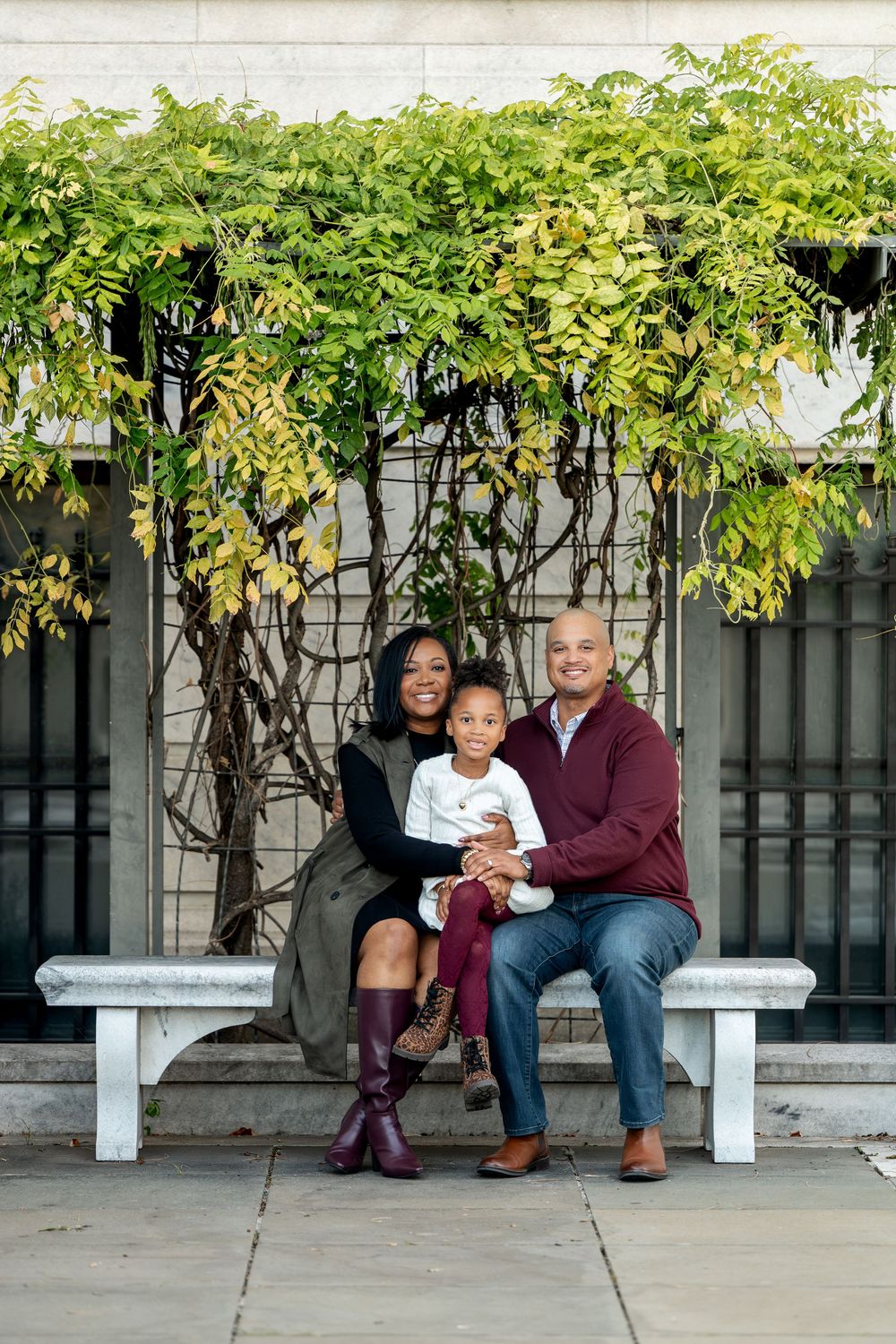 A joyful family of three sits on a bench surrounded by lush green vines on a wall.