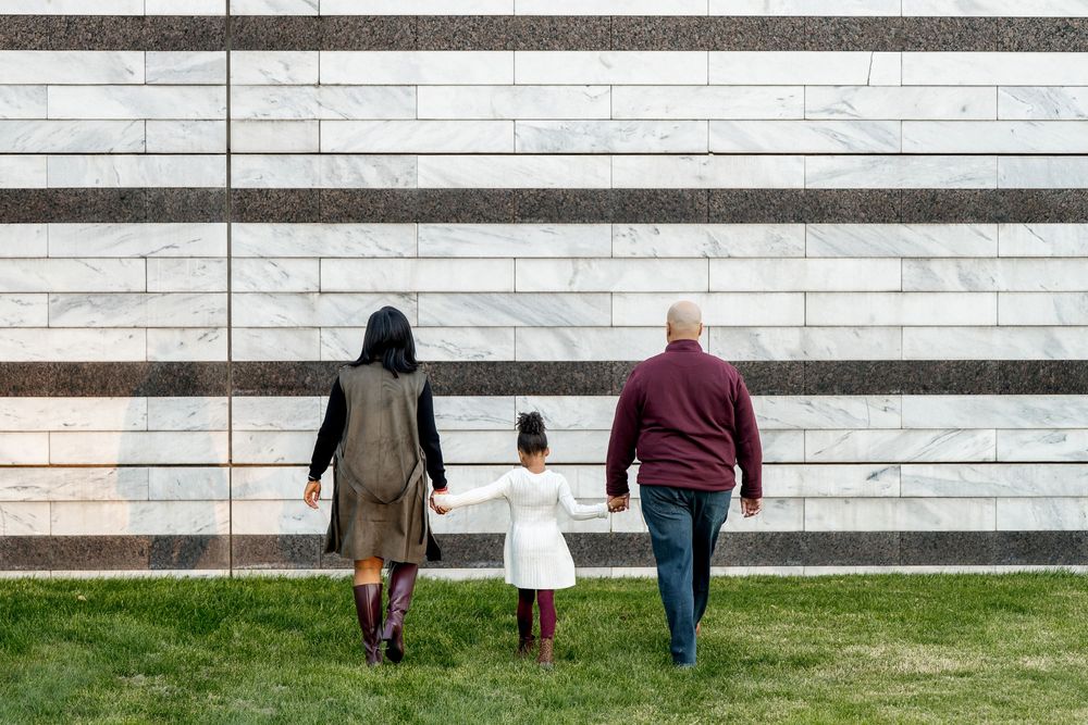 A family of three walks hand in hand across green grass toward a striped marble wall.