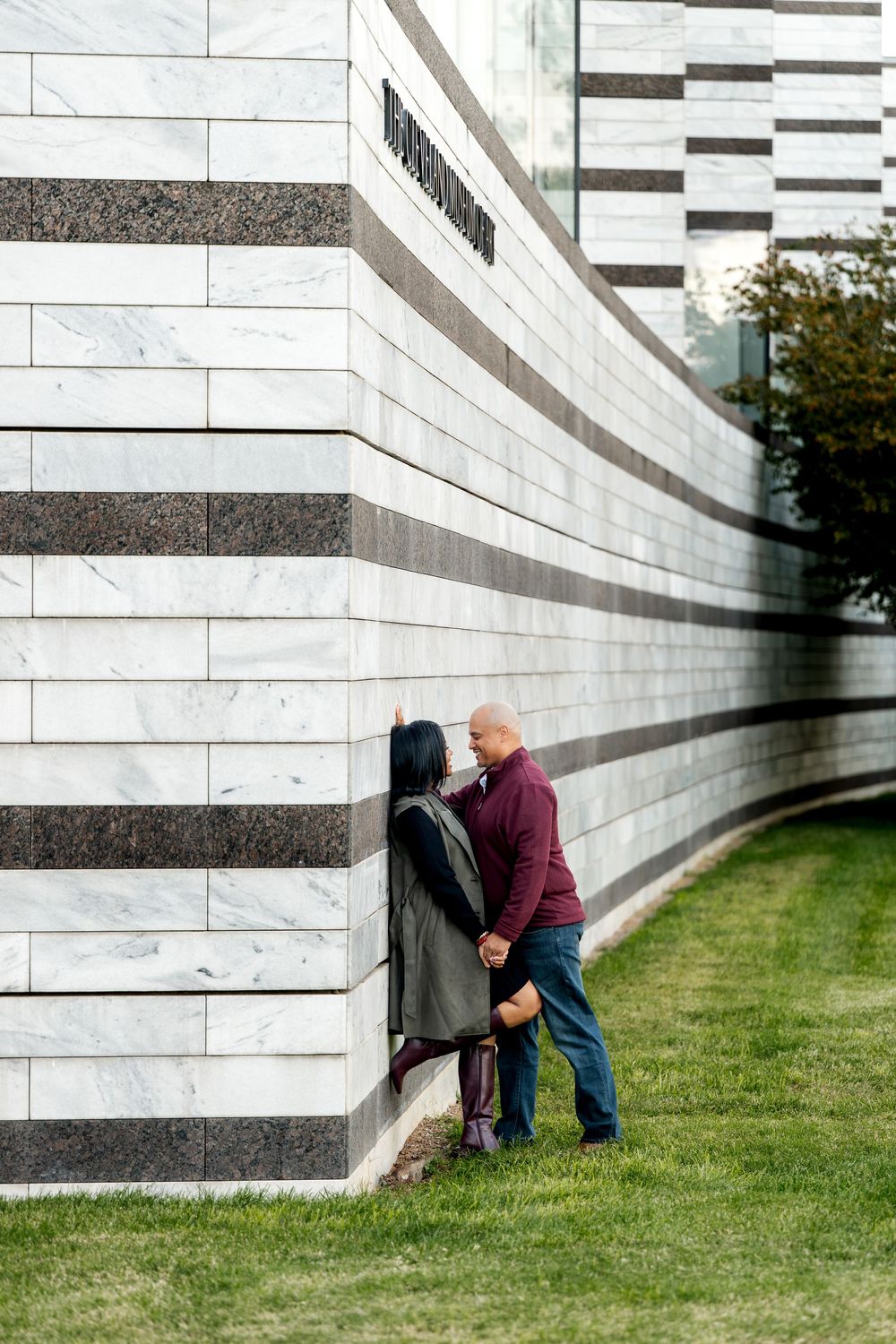 Couple sharing a romantic moment against a striped marble building exterior on a green lawn.