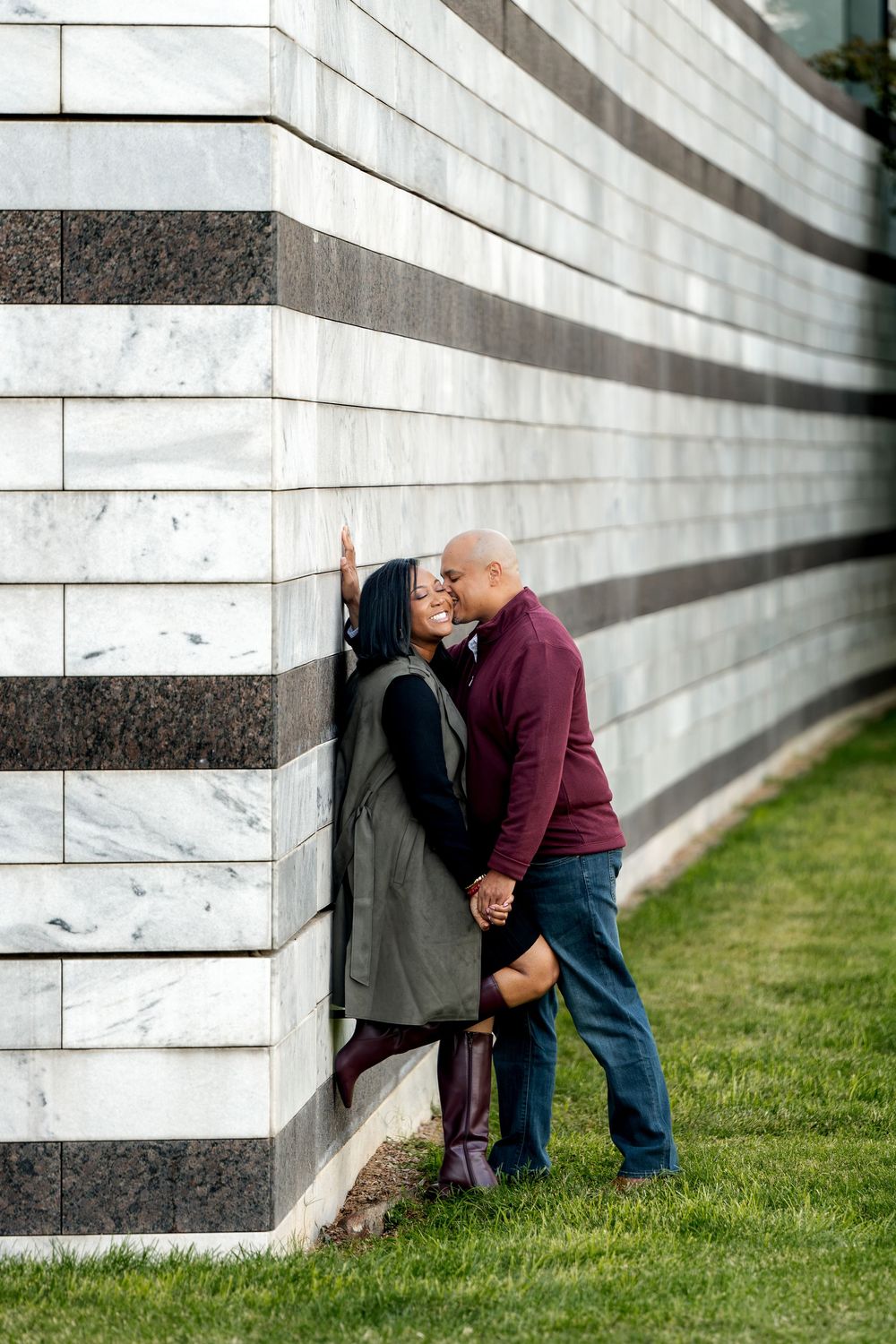 A couple shares a romantic moment leaning against a marble striped wall on a green lawn.
