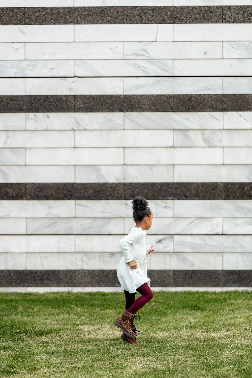 Young girl in white dress and maroon leggings running on grass in front of a striped marble wall.