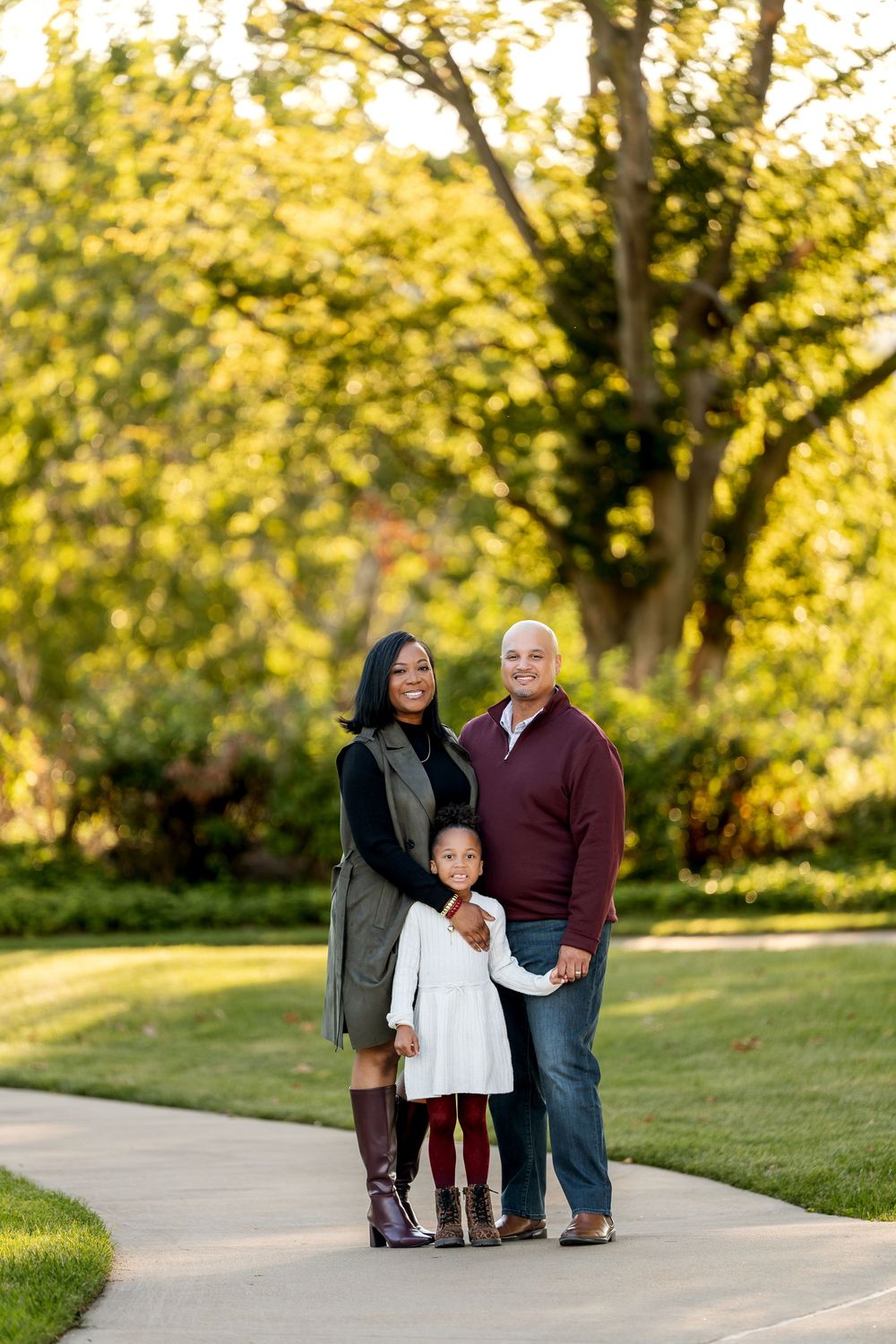 A happy interracial family of three poses on a park path surrounded by lush green autumn trees.