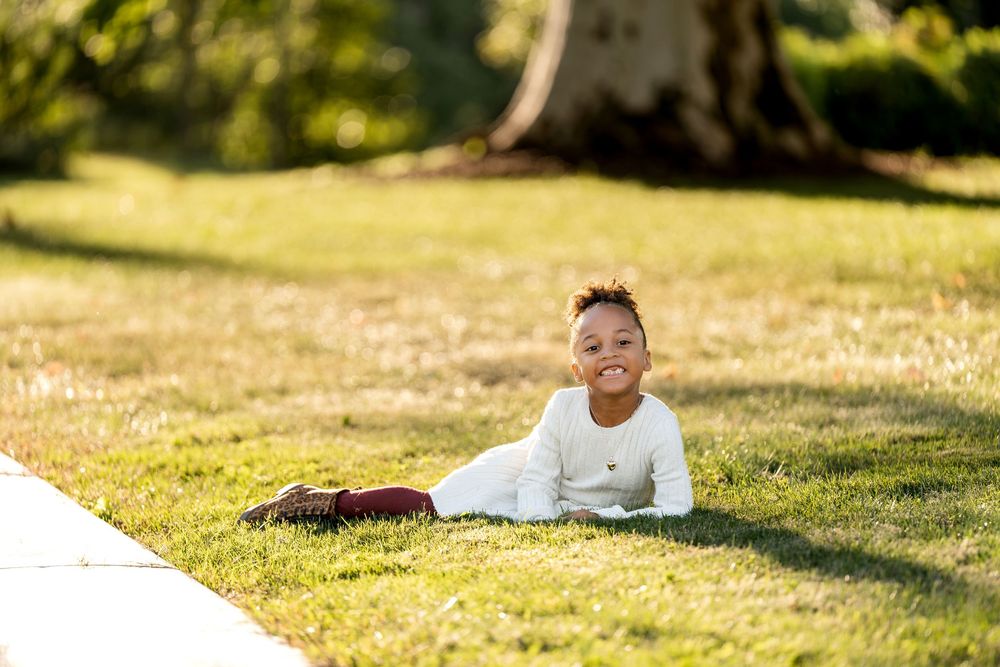 Smiling baby lying on green grass in a sunny park, wearing a white outfit, with trees in the background.