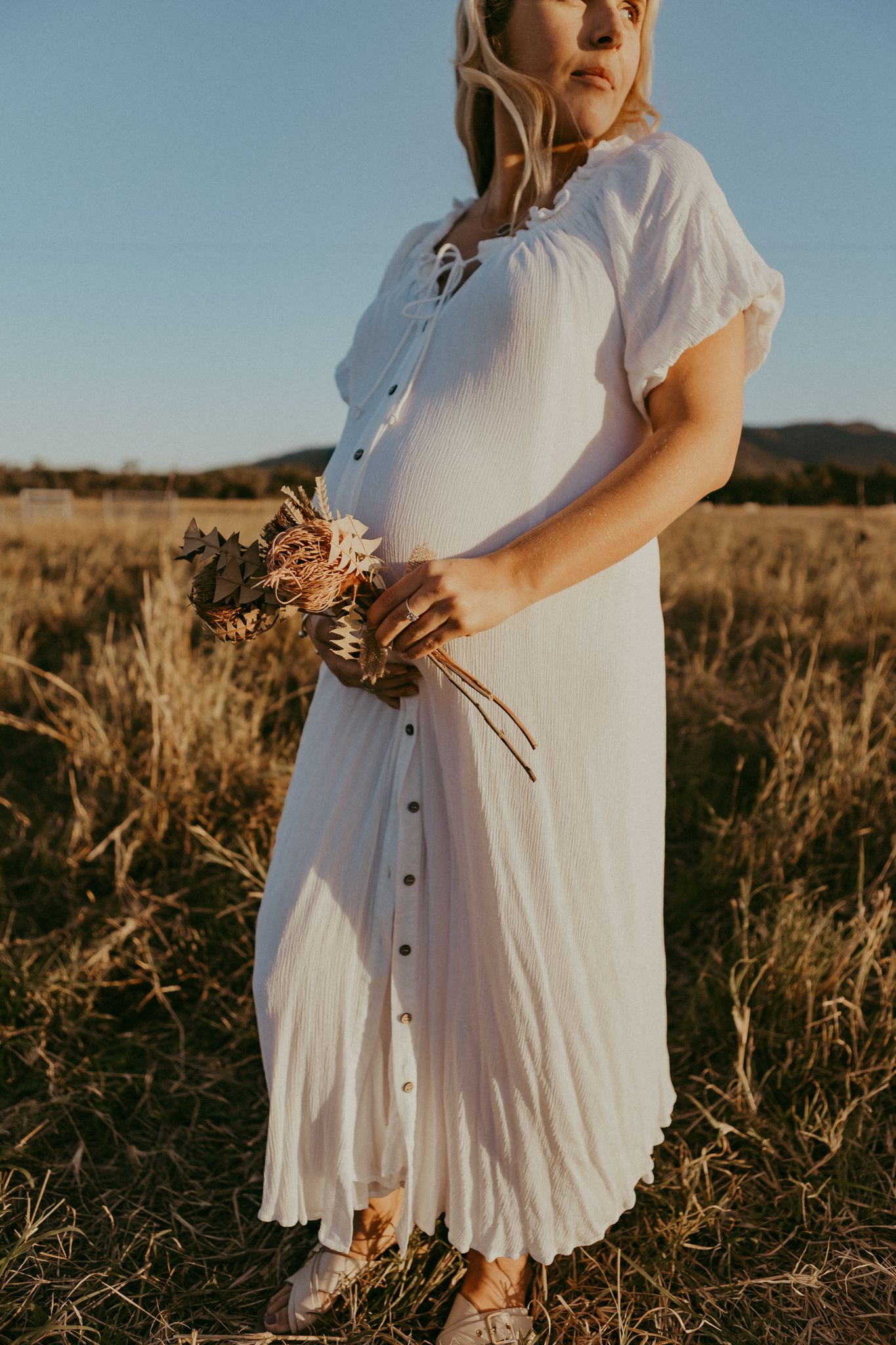 A pregnant woman in a white button-down dress holds dried flowers while standing in a dry field.