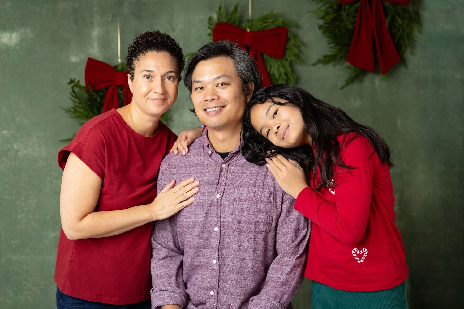 Family of three smiling together in front of holiday wreaths on a green background, wearing festive red outfits.