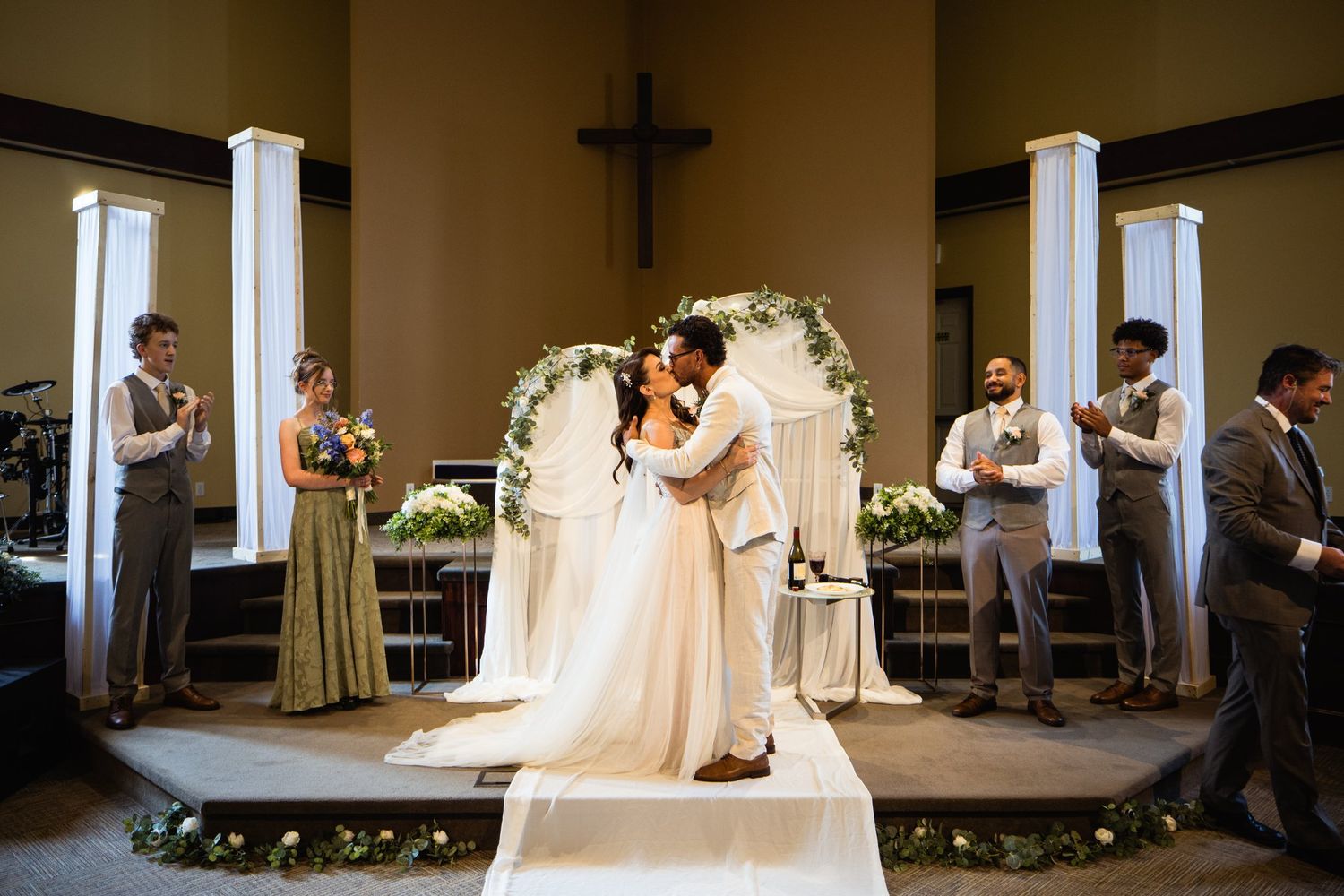 First kiss under a floral arch and wooden cross at King's Gate Church in Cochrane