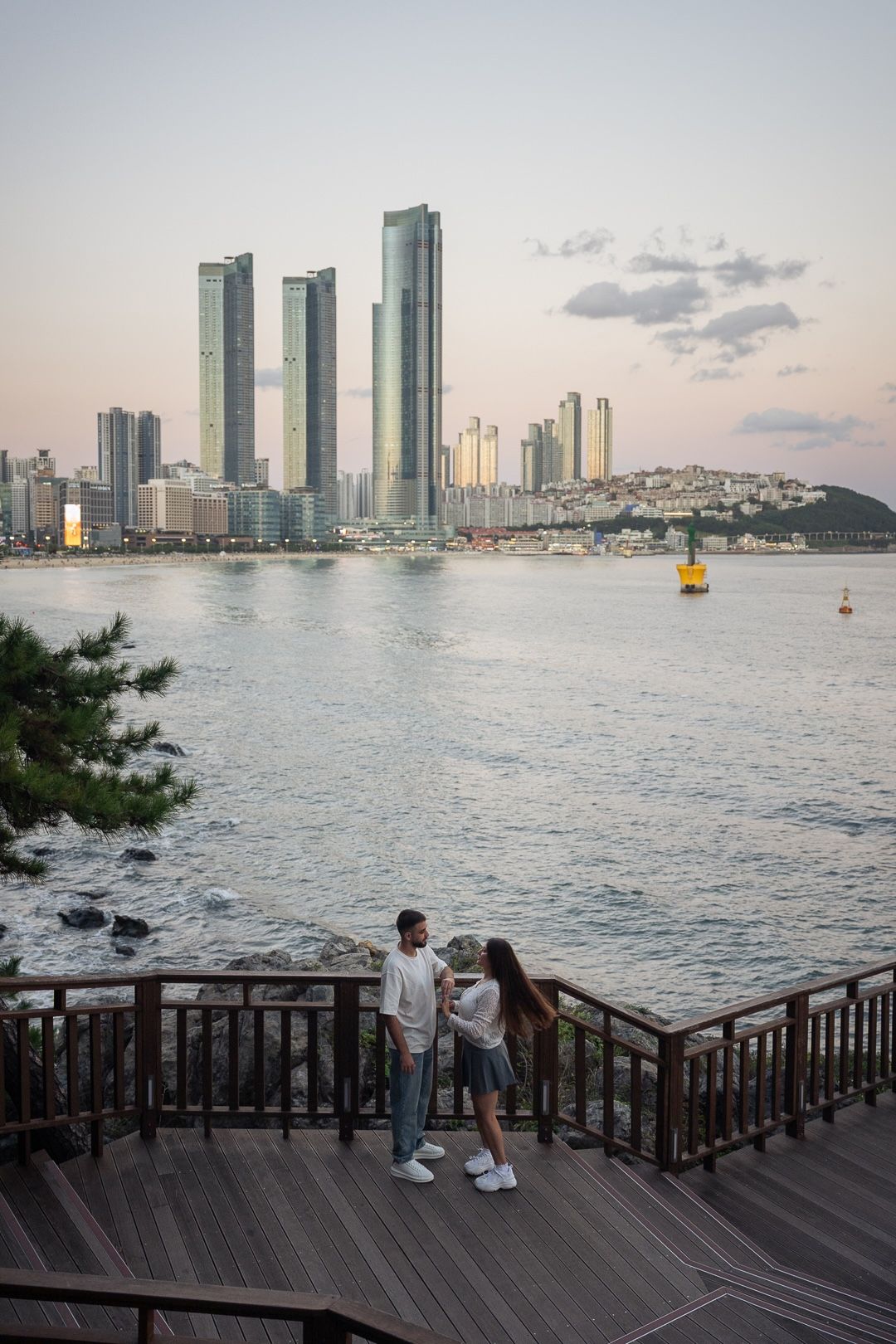 Couple standing on wooden deck overlooking Haeundae Beach skyline with modern skyscrapers at sunset in Busan, South Korea.