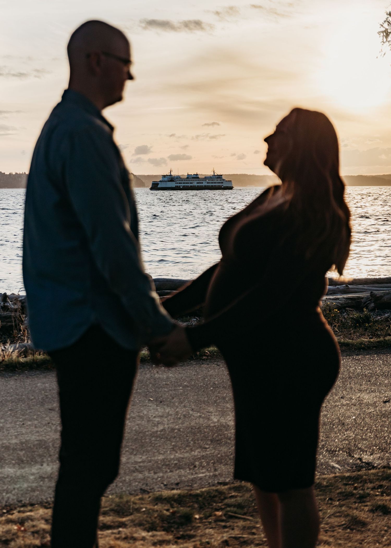 Silhouetted couple holding hands at sunset on a waterfront beach, with a ferry crossing the calm water behind them.