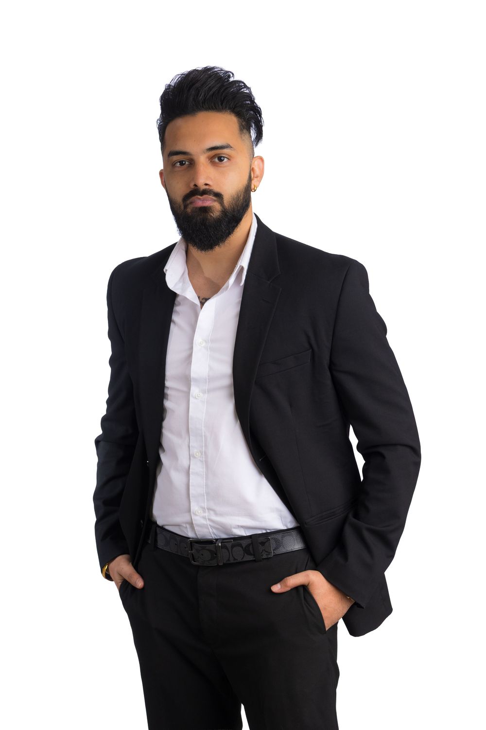 Confident bearded man in a black blazer, white dress shirt and black trousers standing with hands in pockets against a white background.