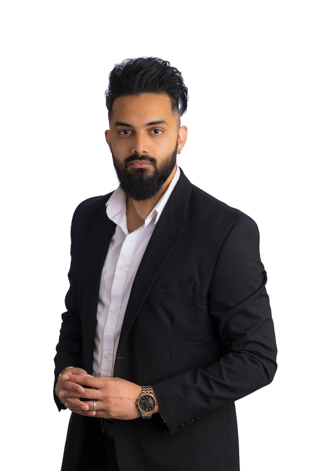 Confident bearded man in a black blazer and white open-collar shirt with a wristwatch, hands clasped, posing against a white background.