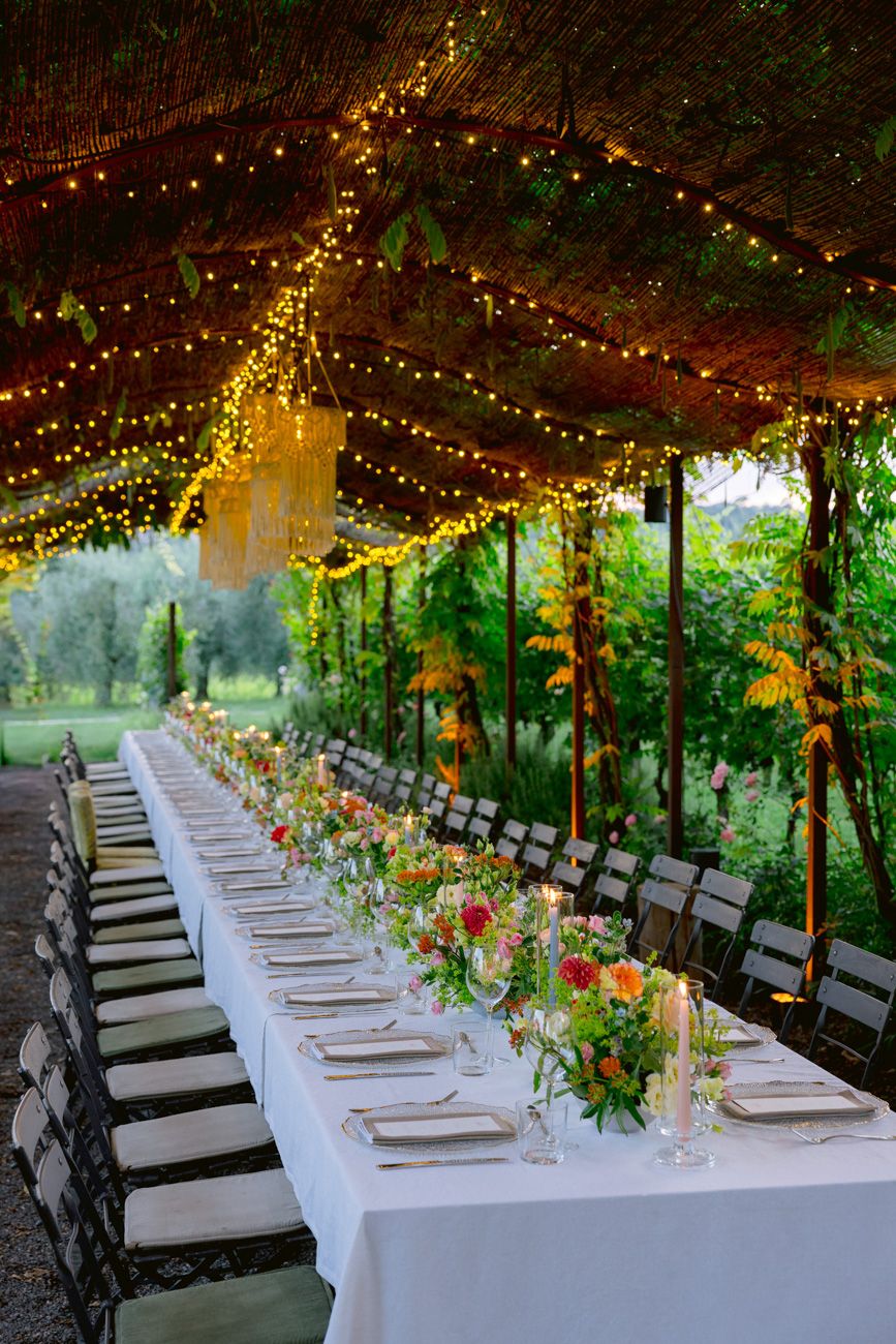 A colourful rehearsal dinner table with candles under a leaf canopy & rope chandeliers in a Lake Garda vineyard. Design by Solidea Events