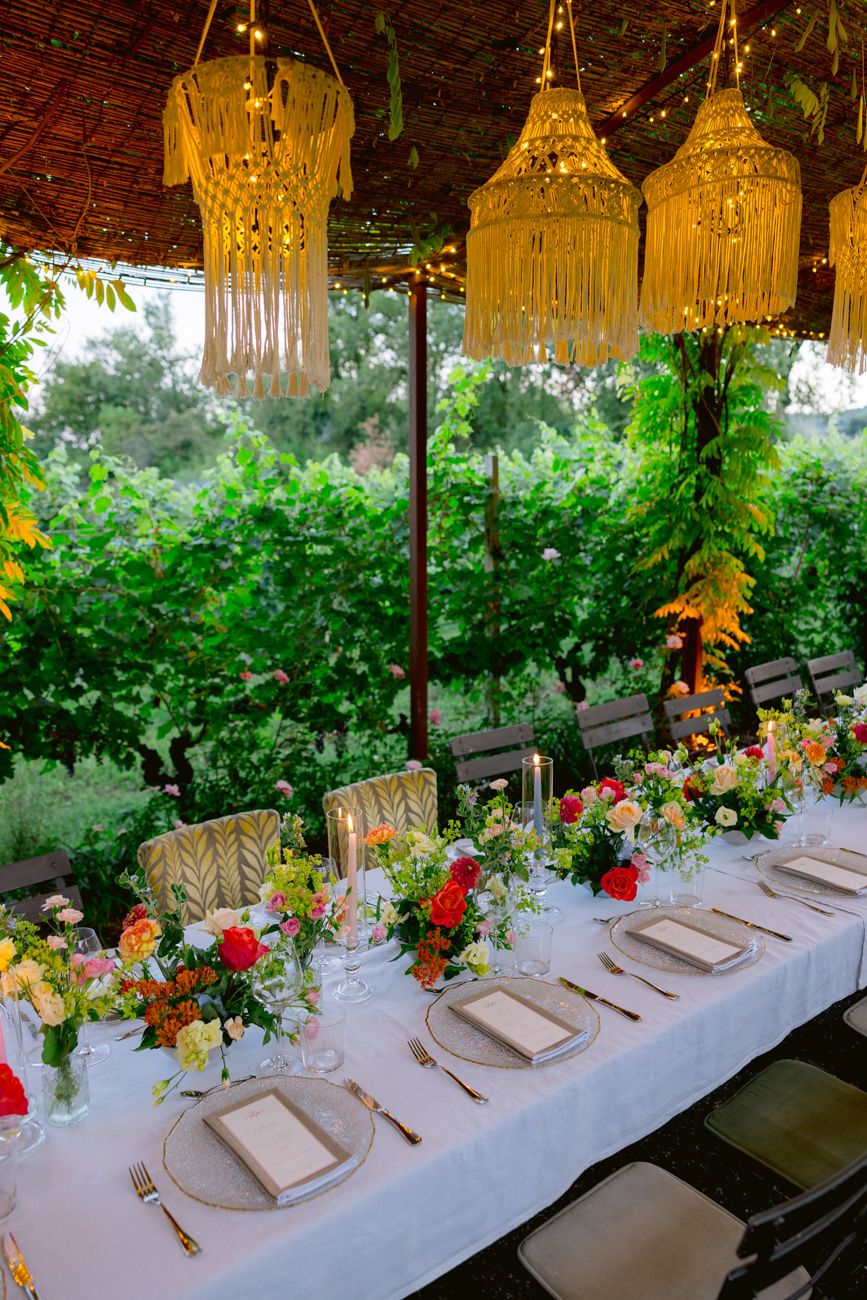 A colourful rehearsal dinner table with candles under a leaf canopy & rope chandeliers in a Lake Garda vineyard. Design by Solidea Events