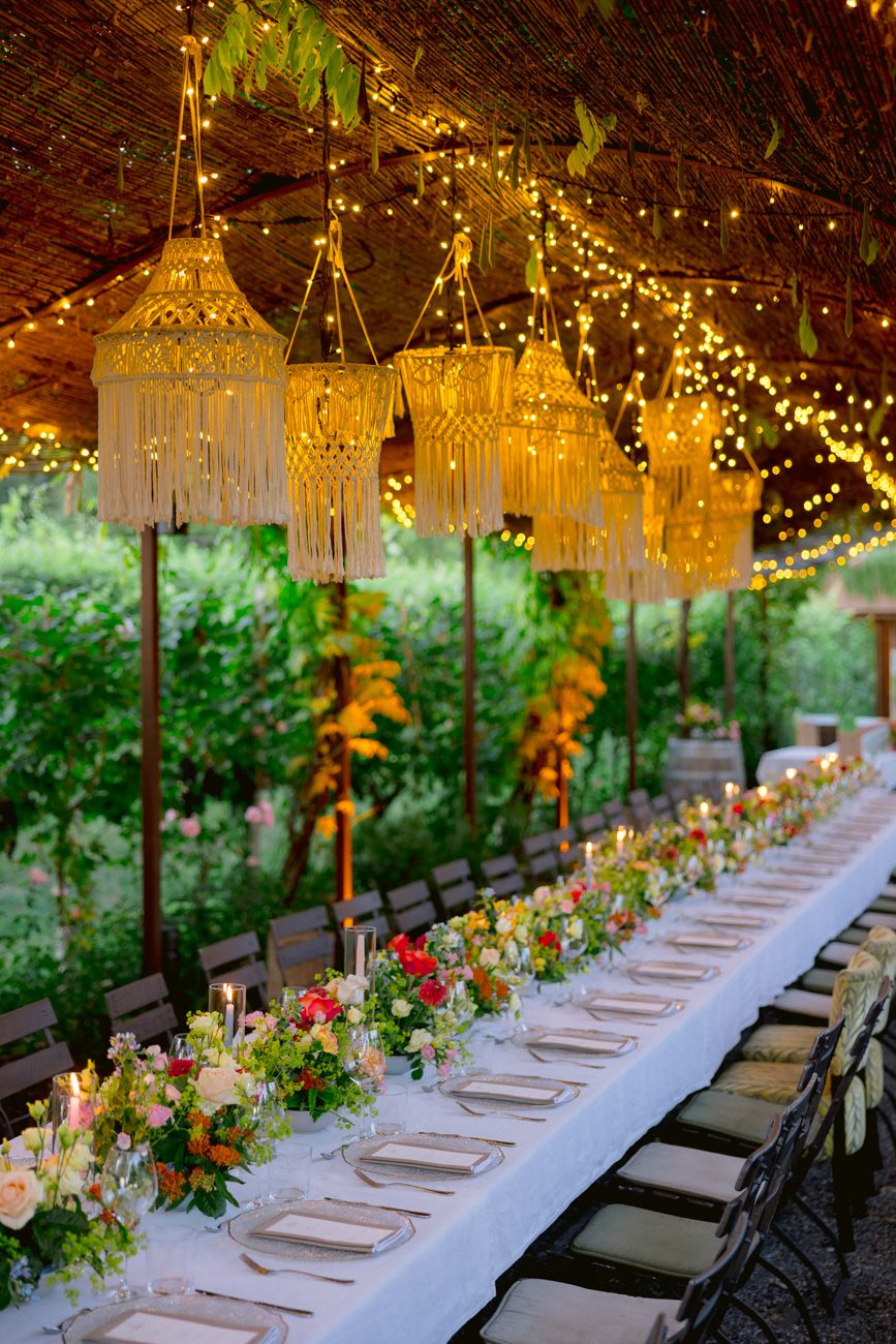 An elegant and colourful rehearsal dinner table with candles under a leaf canopy & rope chandeliers in a Lake Garda vineyard.