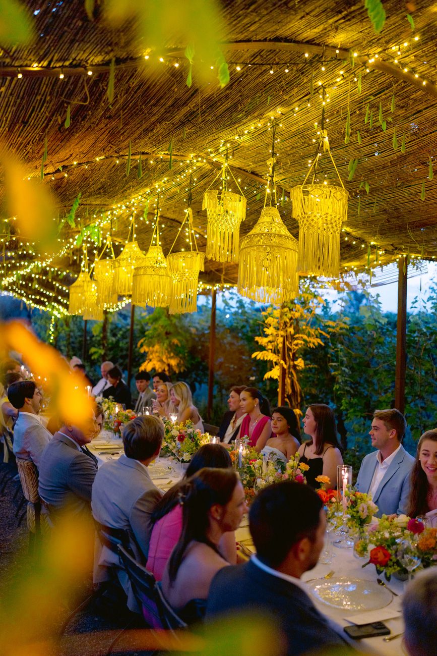 Guests sitting at a rehearsal dinner table under a leaf canopy & rope chandeliers in a Lake Garda vineyard