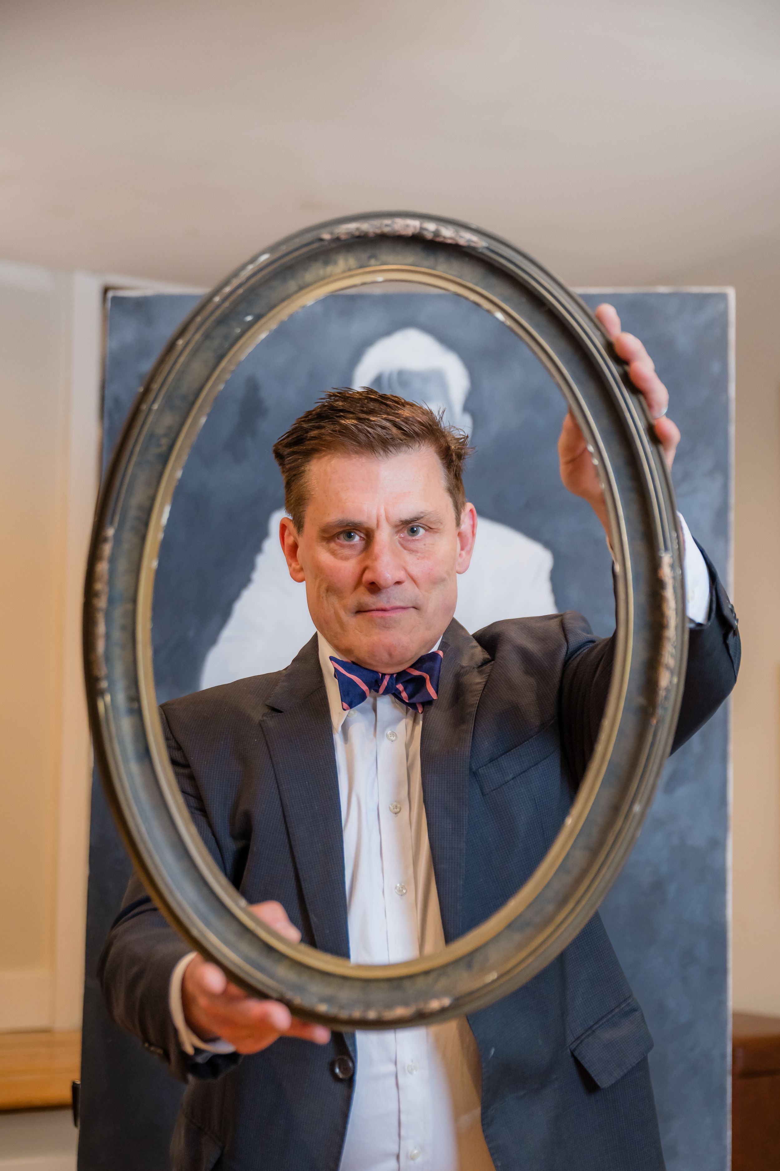 Man in suit and bow tie holds an oval picture frame in front of his face inside an art gallery.