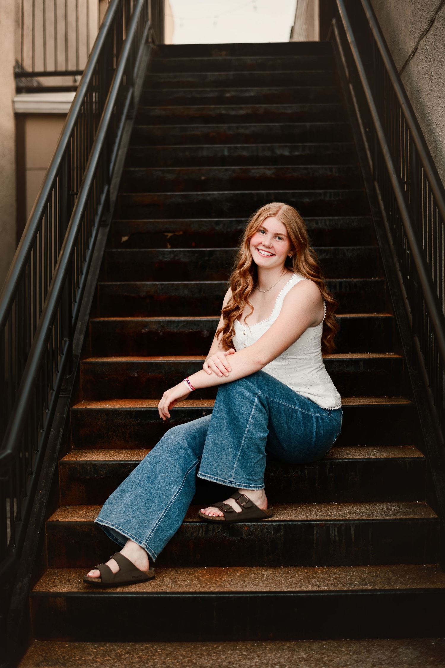 Young woman with long red hair sitting on wooden stairs, wearing white tank top, flare jeans, and black sandals.