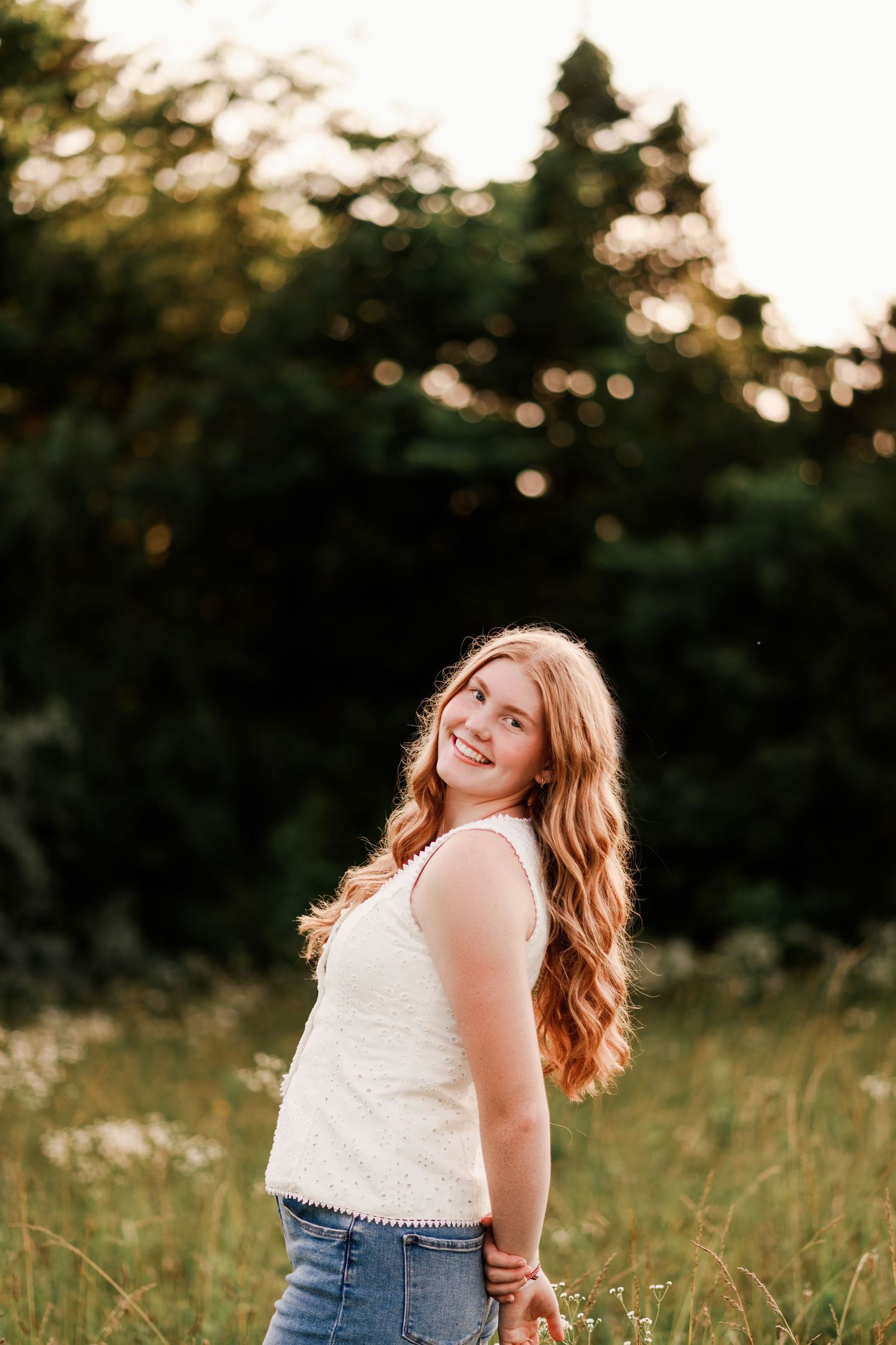Young woman with long red hair smiles in a sunlit field, wearing a white top and jeans, with green trees behind.