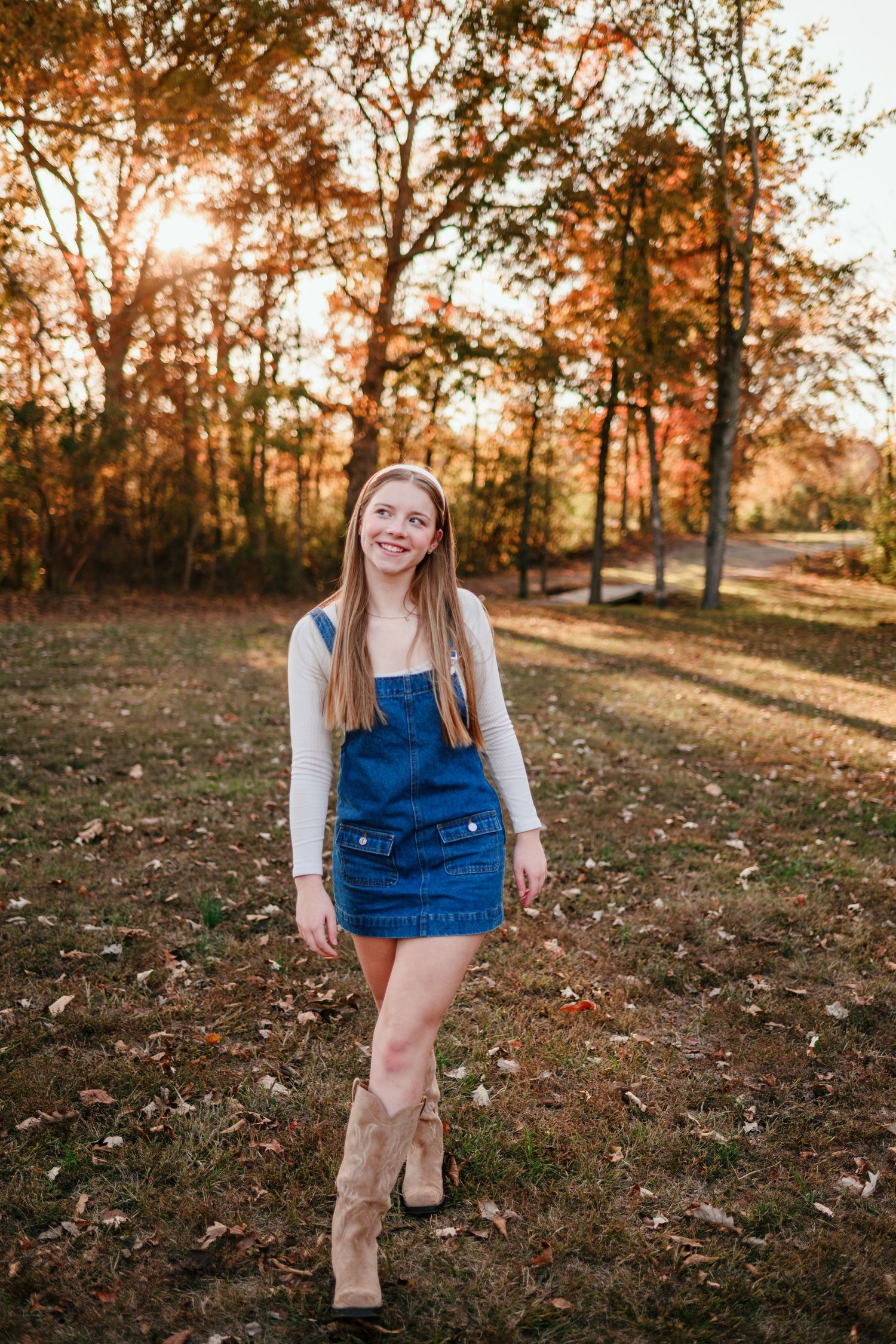 Young woman in denim overalls and cowboy boots smiling in autumn park at golden hour.