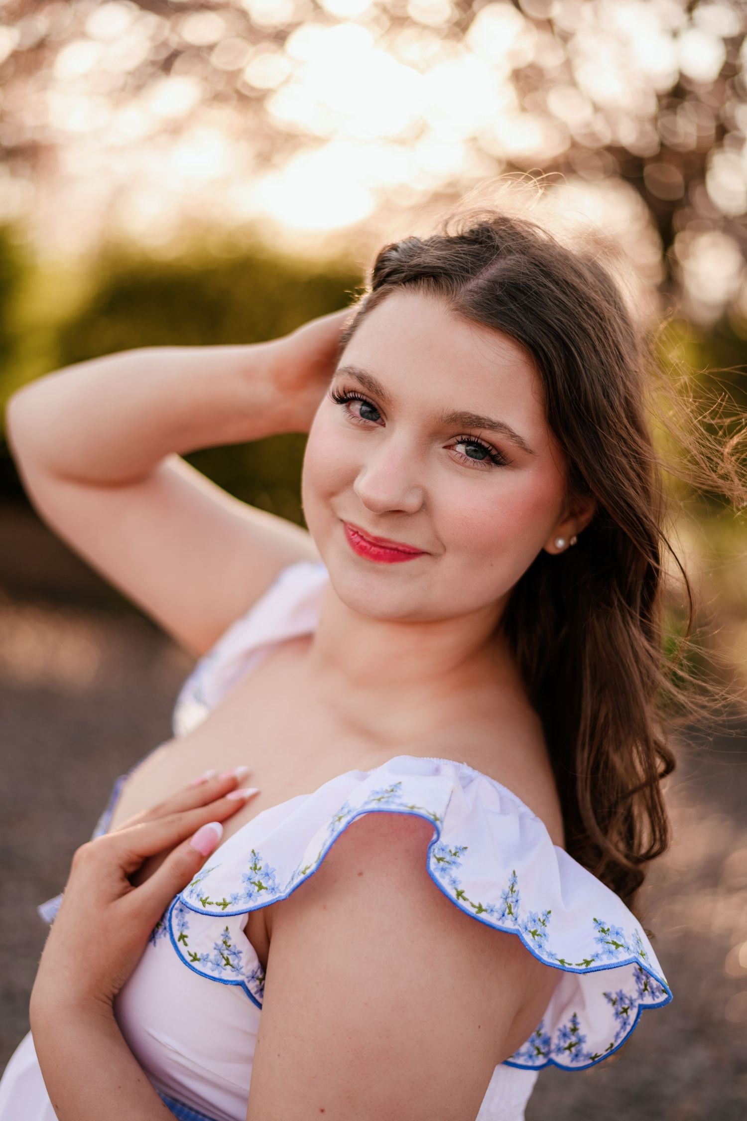 Young woman in off-shoulder white floral dress poses outdoors during golden hour sunset.