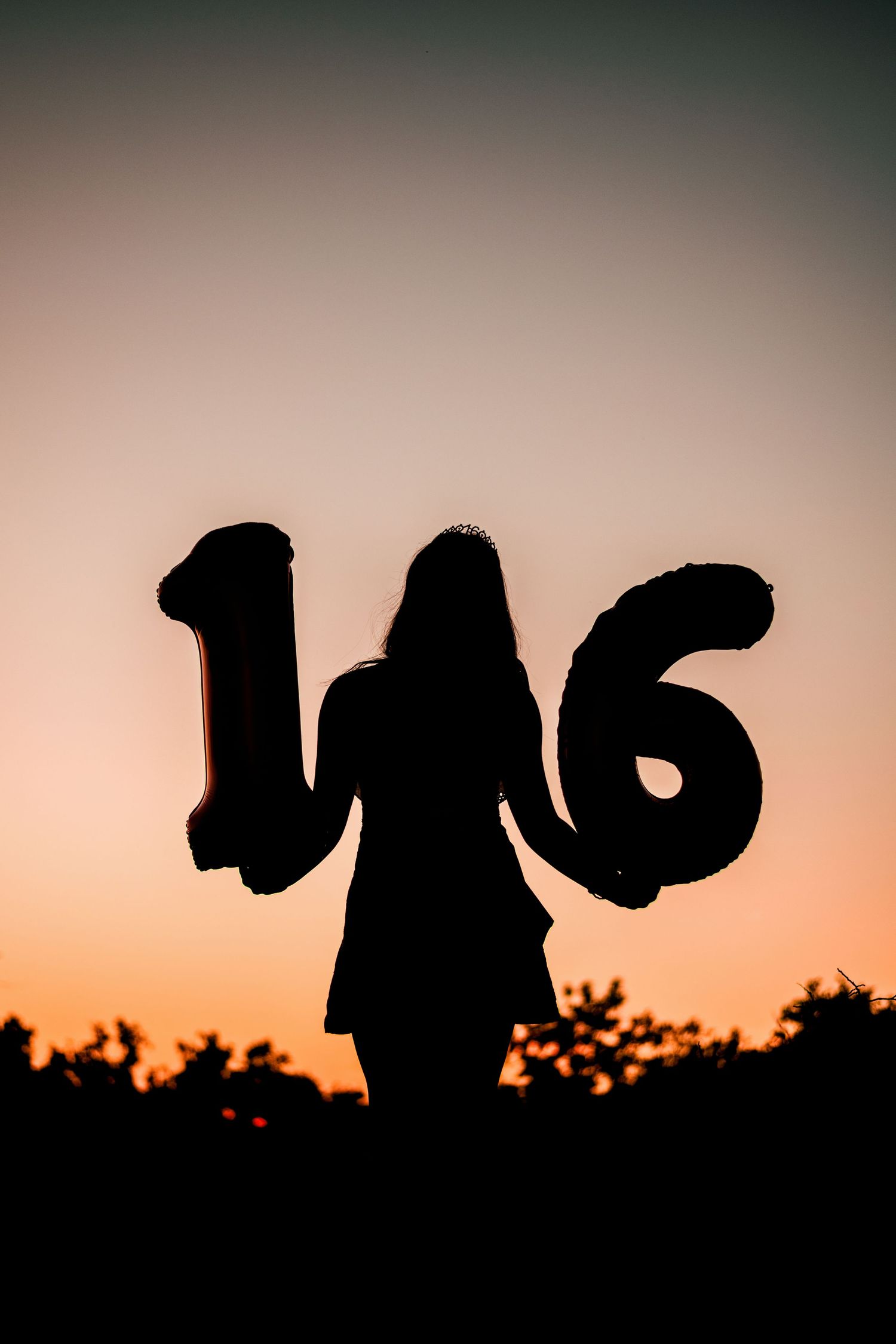 Silhouette of a girl holding number 16 balloons against a golden sunset sky.